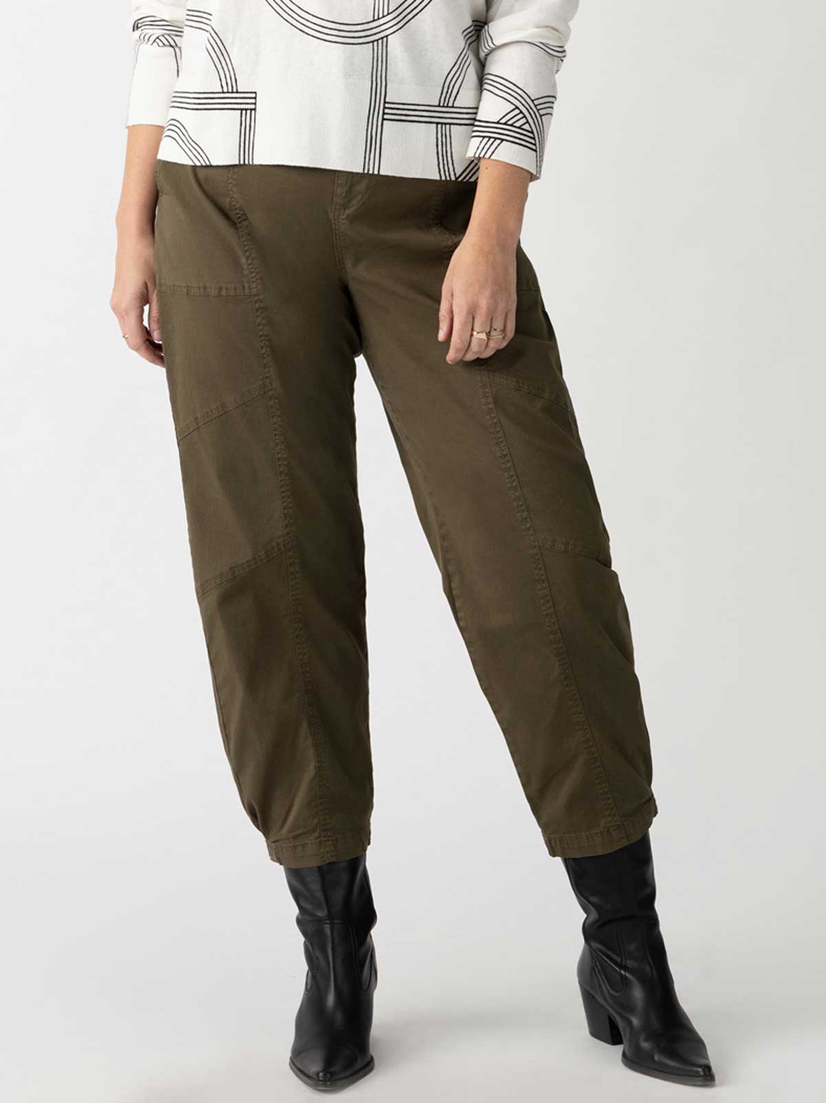 A person wearing the Sahara Tapered Pant in Burnt Olive from Sanctuary Clothing's Inclusive Collection, paired with a white top featuring black geometric patterns and black boots. The image highlights the pants and footwear ensemble.