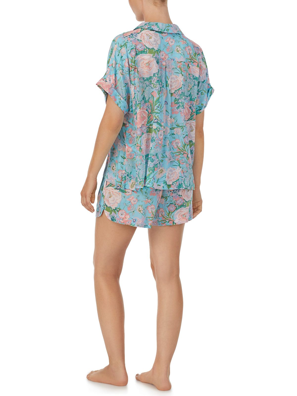 A person stands barefoot wearing the Sanctuary Clothing Short Sleeve Button Up Lounge Set Summer Roses, which features a matching pajama set with a floral print. The set includes a short-sleeved button-up shirt and shorts, both in blue adorned with pink flowers and green leaves. The person is viewed from the back.