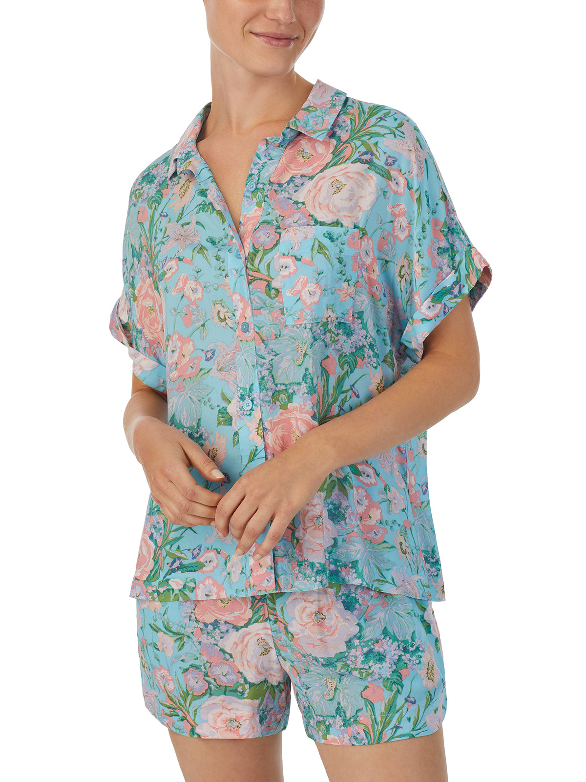 A person is wearing the Short Sleeve Button Up Lounge Set Summer Roses by Sanctuary Clothing, a matching set of light blue pajamas adorned with pink and orange floral patterns. The set comprises a short-sleeve button-up shirt and shorts. The person is smiling, with their hands casually resting at their waist.