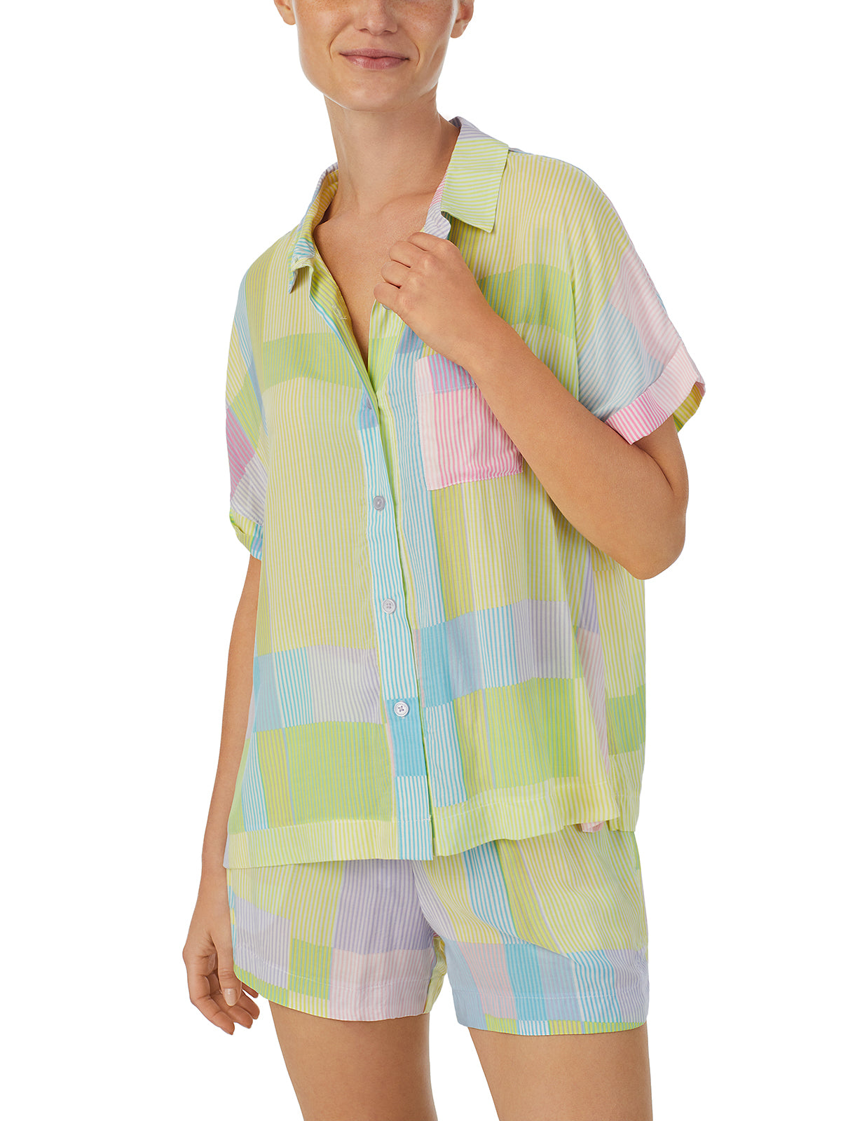 A person is wearing the Sanctuary Clothing Short Sleeve Button Up Lounge Set Multi Stripe, comprising a shirt and matching shorts. The outfit features a pastel color block pattern with shades of yellow, pink, blue, and green. The individual is smiling and has their hand near the collar of the shirt.