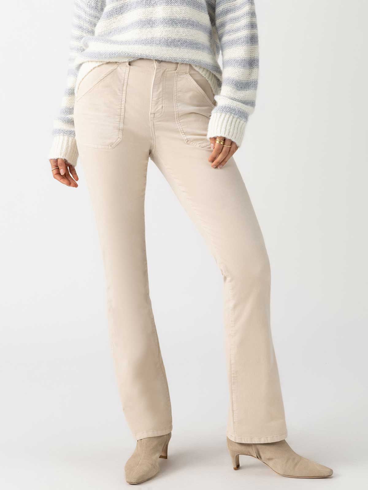 A person wearing beige velveteen Hayden bootcut pants from Sanctuary Clothing, a light gray and white striped sweater, and toasted almond ankle boots. The image is focused on the lower half of the body, with hands partially visible in the sweater sleeves.