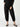 A person is standing, dressed in a black long-sleeve top and the "Sahara Tapered Pant Black" from the Inclusive Collection by Sanctuary Clothing. Their hands are in their pockets, and they are wearing black ballet flats. The background is a plain, light color.