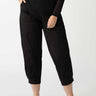 A person is standing, dressed in a black long-sleeve top and the "Sahara Tapered Pant Black" from the Inclusive Collection by Sanctuary Clothing. Their hands are in their pockets, and they are wearing black ballet flats. The background is a plain, light color.