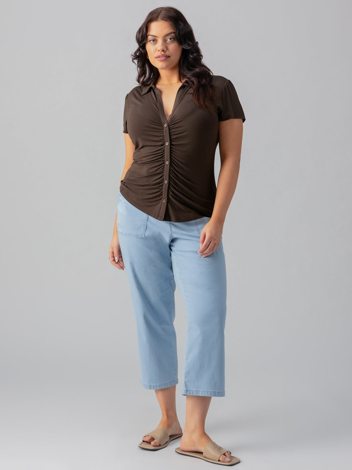 A woman with long, wavy hair is standing against a plain background. She is wearing the Dream Button Up Mud Bath top from Sanctuary Clothing's Inclusive Collection, light blue cropped jeans, and light-colored open-toed sandals. She is looking directly at the camera.