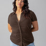 A person with long, wavy hair is wearing the Dream Button Up Mud Bath Inclusive Collection blouse from Sanctuary Clothing. The brown short-sleeved button-up blouse features a ruched front detail and is paired with light blue jeans. They stand against a plain grey background, looking at the camera with a neutral expression.