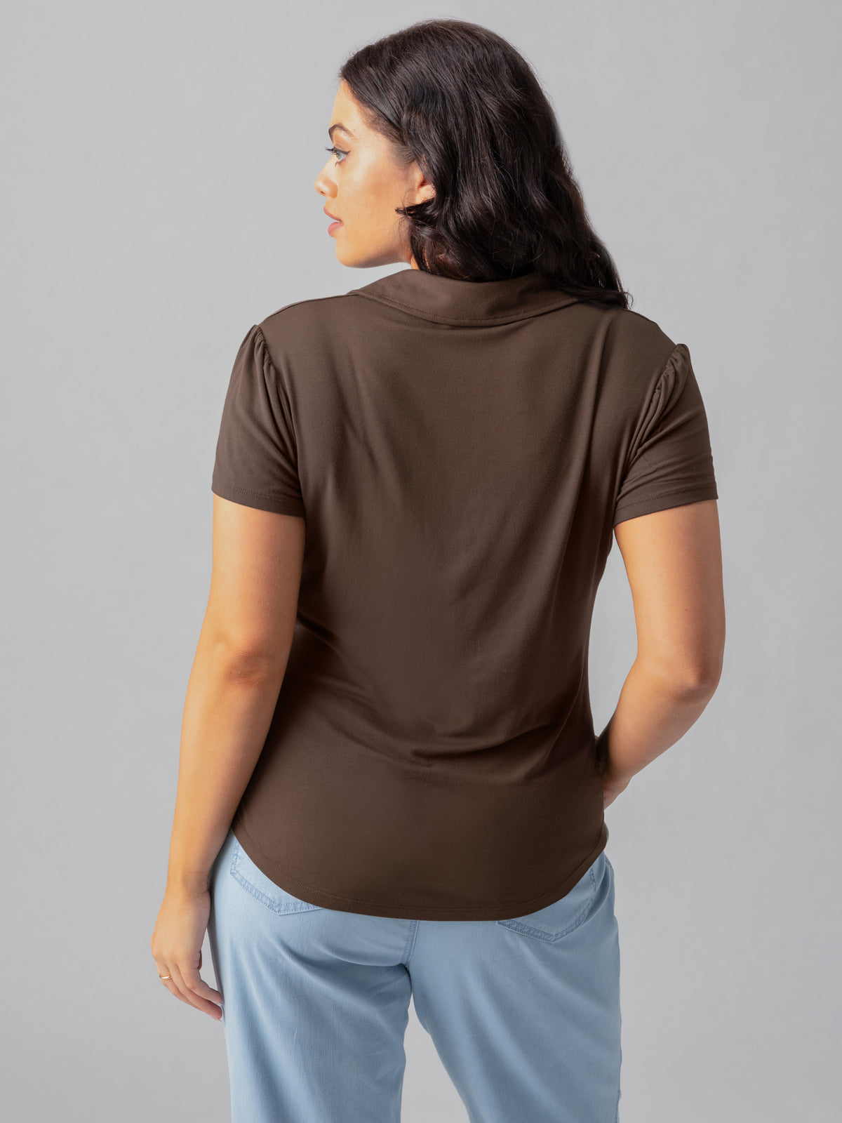 A woman with dark hair is facing away from the camera, partially turned to the side. She is wearing the Dream Button Up Mud Bath inclusive collection short-sleeved brown top by Sanctuary Clothing and light blue pants. The background is plain and gray.