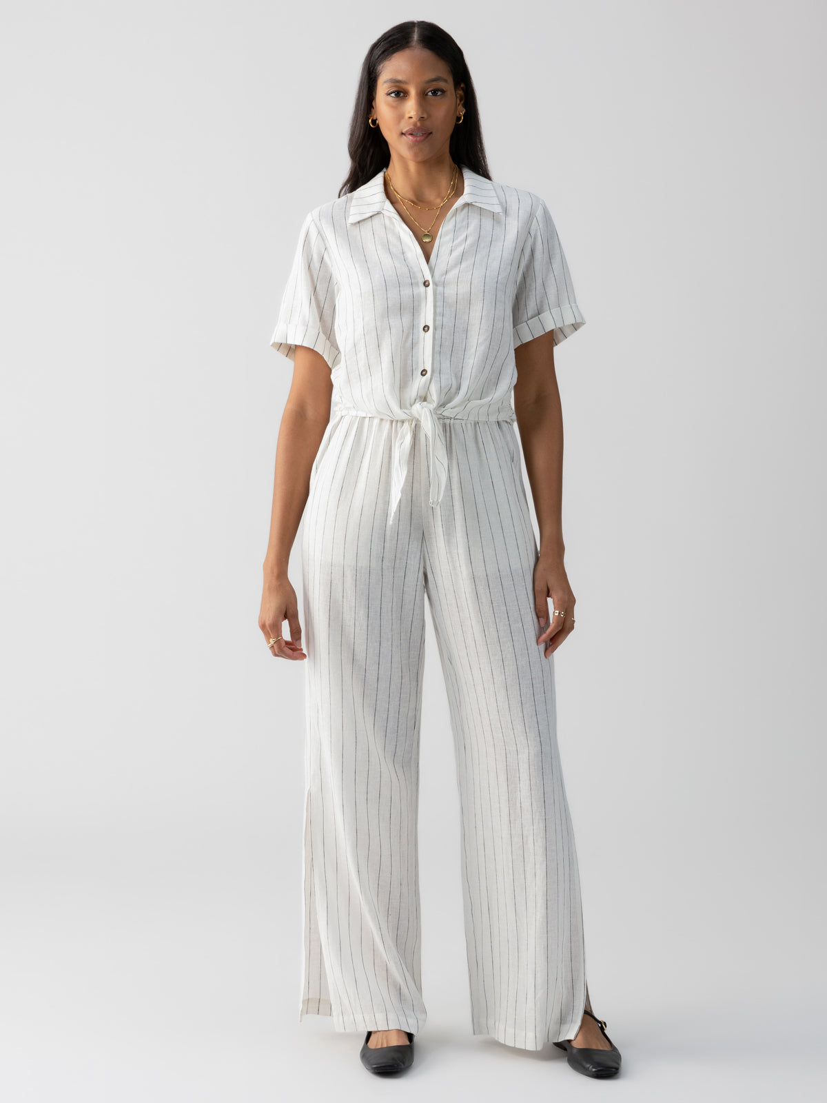 A woman stands against a plain background wearing the Ocean Shirt Biarritz Stripe by Sanctuary Clothing, a white button-up jumpsuit with thin vertical stripes and a tie waist. She has long hair and accessorizes with earrings, a necklace, and black flats. She appears to be looking directly at the camera.