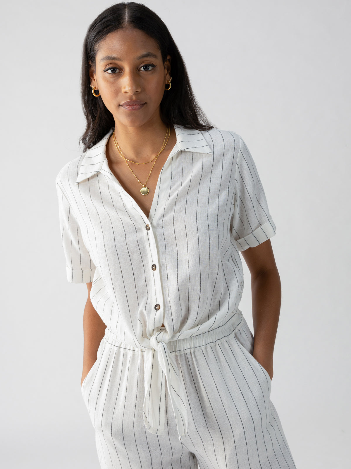 A person with long dark hair is wearing Sanctuary Clothing's Ocean Shirt Biarritz Stripe, a light-colored, vertically striped jumpsuit with short sleeves and a tie around the waist. They are accessorized with gold necklaces and hoop earrings. Their hands are casually in their pockets as they stand against a plain background.