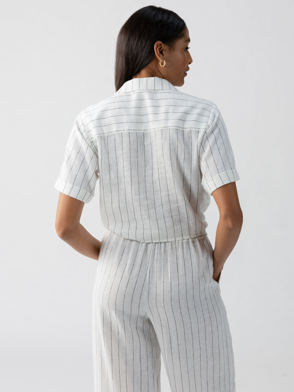 A woman with long hair stands facing away, wearing the Ocean Shirt Biarritz Stripe from Sanctuary Clothing. The short-sleeved, collared shirt is paired with matching high-waisted pants, both featuring vertical pinstripes. The light-colored outfit showcases her hands in her pockets against a plain, light-colored background.