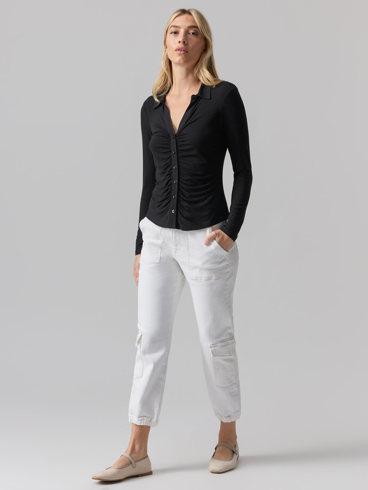 A woman with long blonde hair stands against a light gray background. She is wearing the Dreamgirl Button Up Black from Sanctuary Clothing, along with white cropped trousers with pockets and beige flat shoes. She has a relaxed and confident posture.