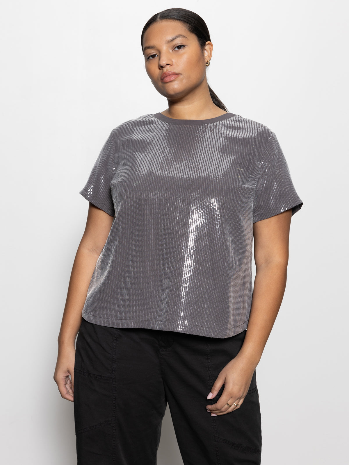 A person with dark hair tied back wears the Sanctuary Clothing's "Perfect Sequin Tee Gunmetal" from their Inclusive Collection, paired with black pants, standing against a plain white background. Their expression is neutral, and their hands rest by their sides.