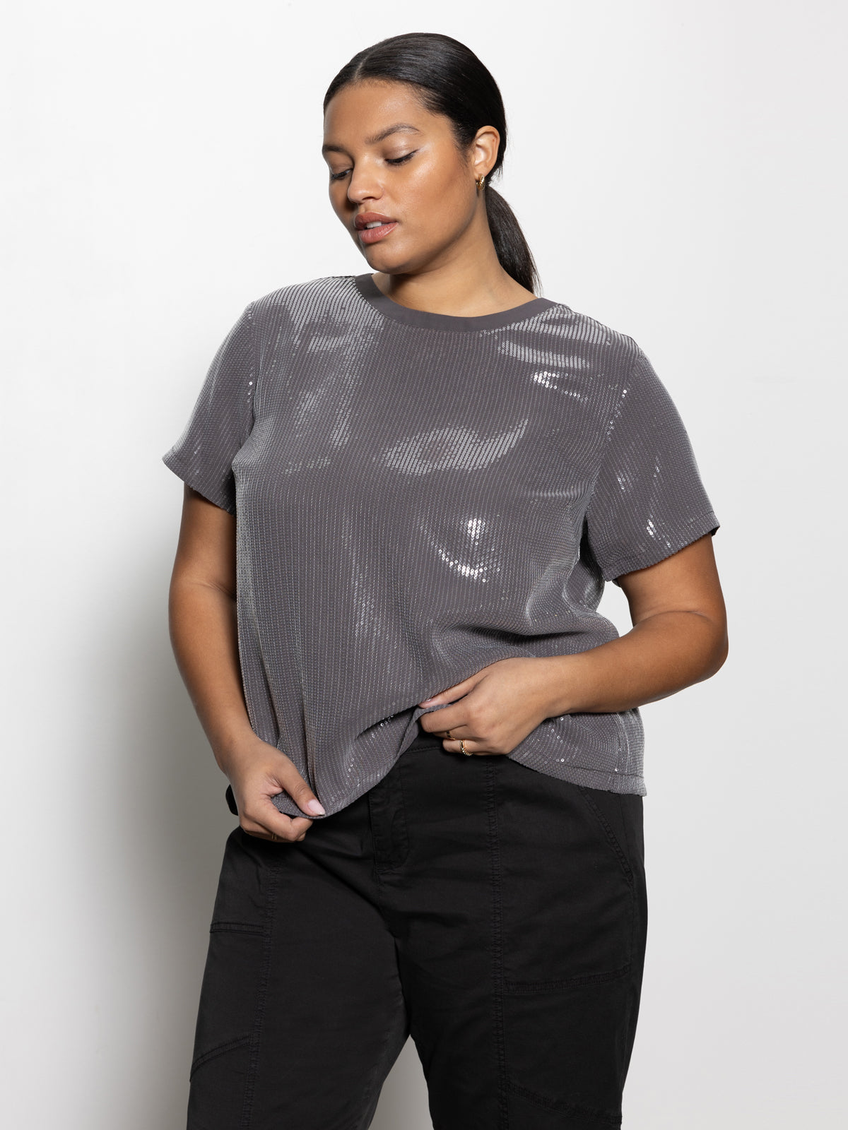 A person wearing Sanctuary Clothing's "perfect sequin tee gunmetal" from the inclusive collection pairs it with black pants, standing against a plain white background. They gaze downwards while adjusting the shirt with one hand. Their hair is styled in a low ponytail.