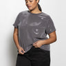 A person wearing Sanctuary Clothing's "perfect sequin tee gunmetal" from the inclusive collection pairs it with black pants, standing against a plain white background. They gaze downwards while adjusting the shirt with one hand. Their hair is styled in a low ponytail.