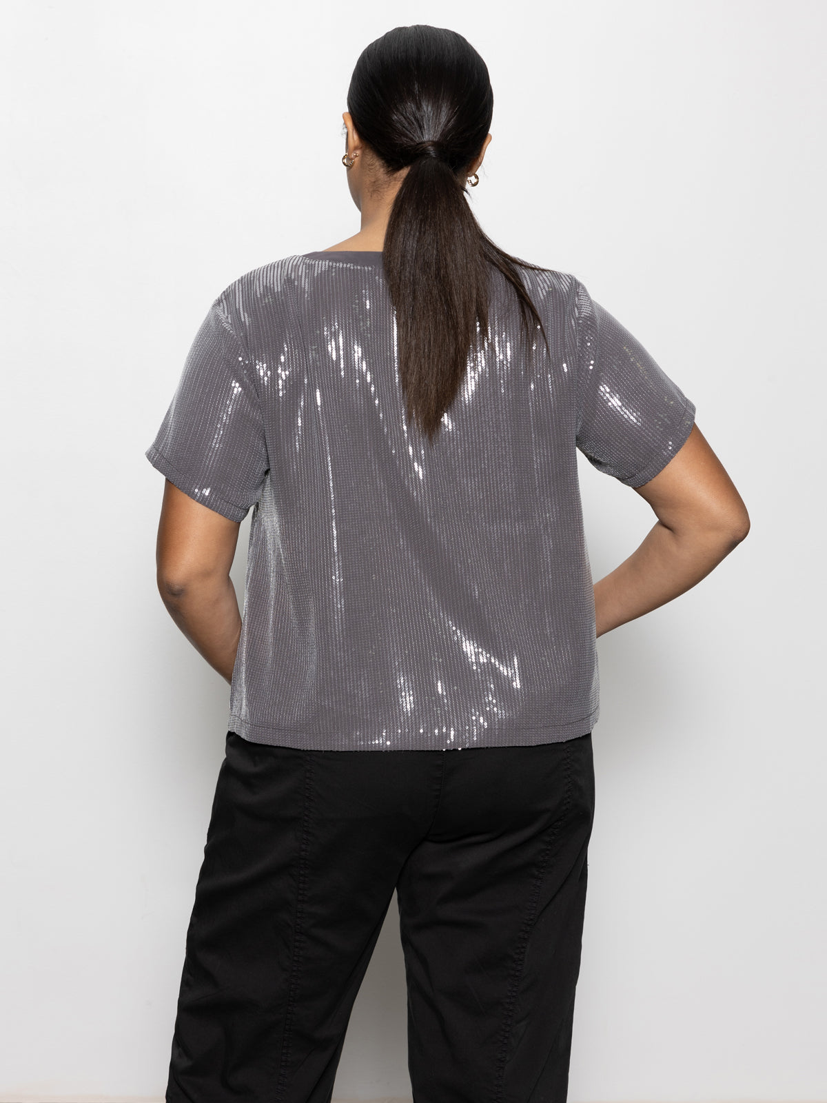 A person with long, dark hair tied in a ponytail is facing away, wearing the Sanctuary Clothing "Perfect Sequin Tee" in gunmetal from the Inclusive Collection and black pants against a plain white background.