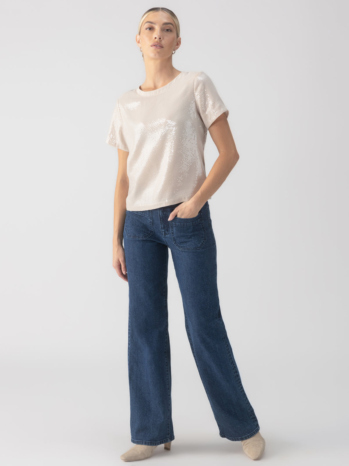 A person stands against a neutral background, dressed in the Perfect Sequin Tee Frosted Almond by Sanctuary Clothing and a pair of blue wide-leg jeans. They have their left hand in their pocket and are wearing beige shoes. Their short hair is slicked back with a headband.