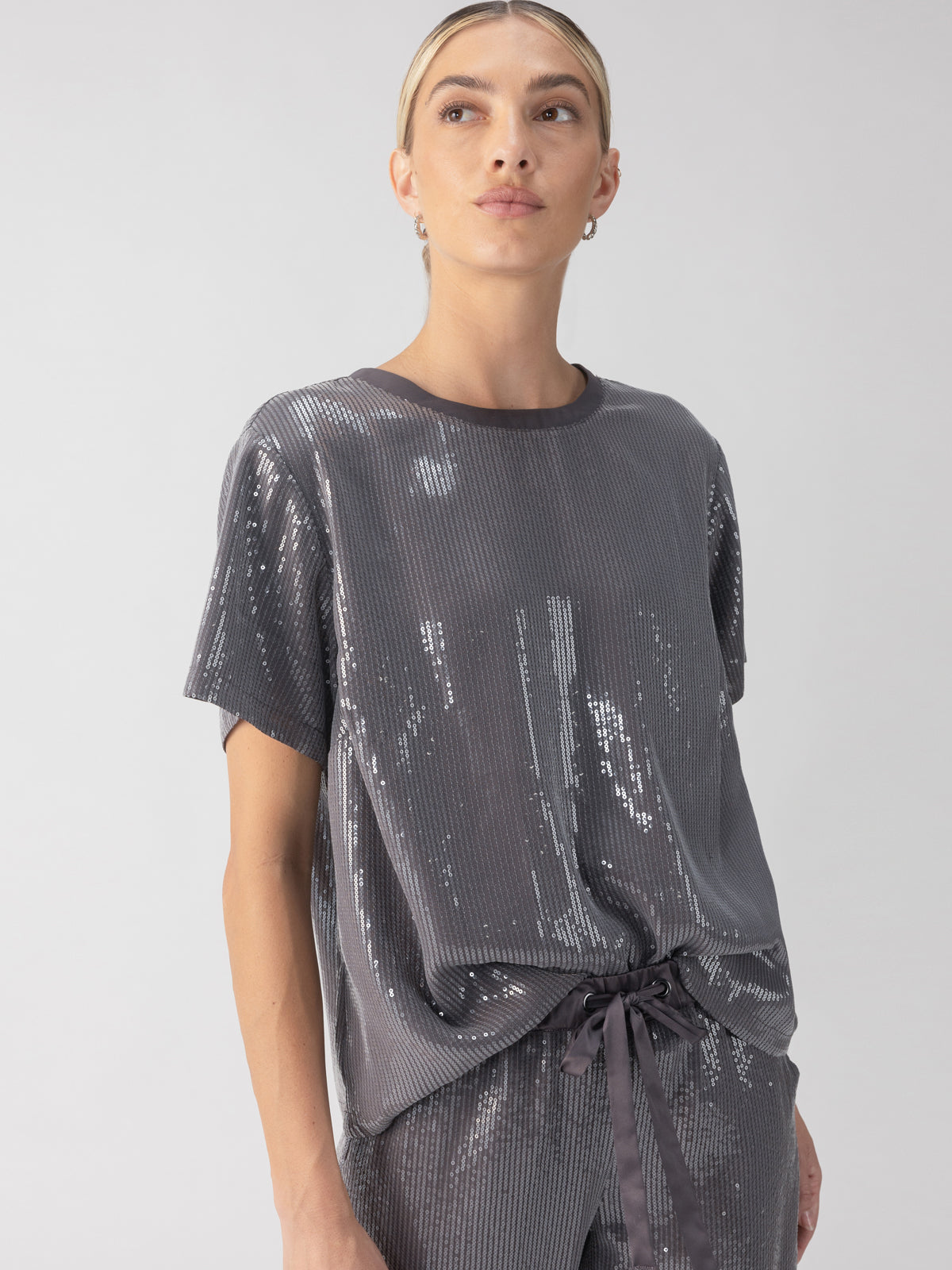 A person wearing the Perfect Sequin Tee Gunmetal by Sanctuary Clothing stands against a plain background. This top features a shiny, silver-gray sequin design with a loose fit and tied hem, beautifully reflecting light to create a shimmering effect. The individual has short hair and gazes slightly to the side.