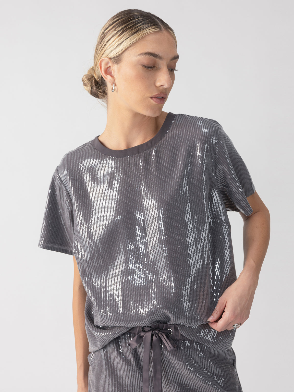 A person dressed in the Sanctuary Clothing Perfect Sequin Tee Gunmetal, which sparkles in various shades of gray and has short sleeves, is paired with matching pants. Their hair is styled neatly in a bun, and they are posing slightly to the side against a simple, light gray background.