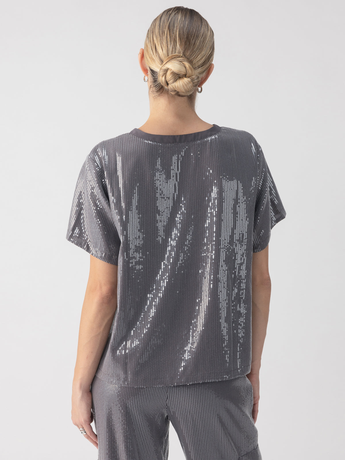 A person with light brown hair in a neat bun wears the Sanctuary Clothing Perfect Sequin Tee Gunmetal, along with matching pants featuring vertical reflective stripes. The person is facing away against a plain background, showcasing the short sleeves and relaxed fit of the outfit.