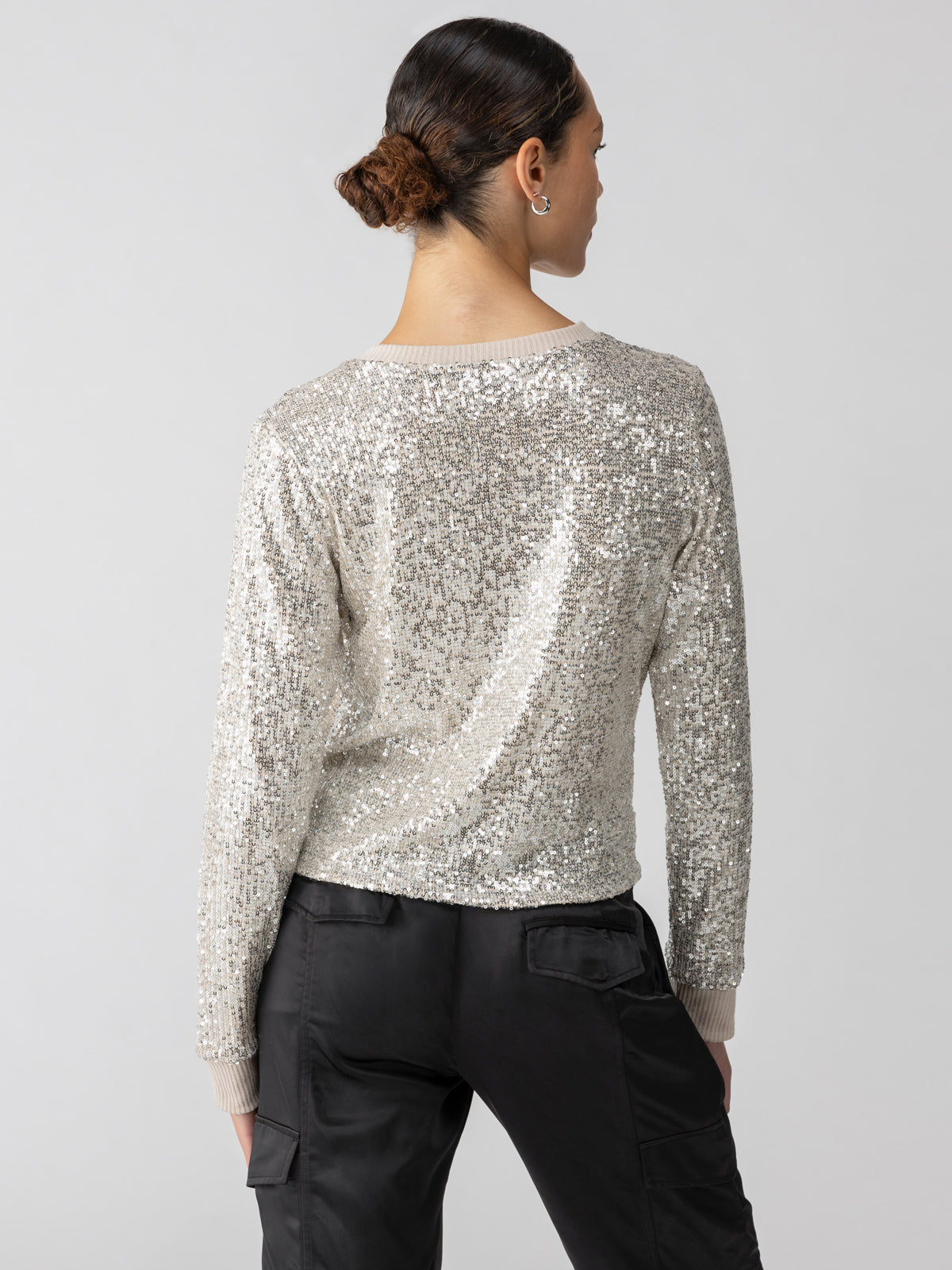 A person with a neat bun hairstyle is facing away from the camera, showcasing the back of the Sparkle Together Top Champagne by Sanctuary Clothing—a silver sequined long-sleeve top paired with dark trousers featuring pockets on the legs. The background is neutral and plain.