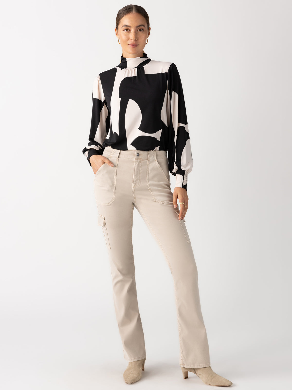 A woman stands against a neutral background, wearing the On The Spot Blouse Formation by Sanctuary Clothing paired with beige trousers. Her hair is tied back, and she has her hands in her pants pockets, exuding a relaxed and confident posture.