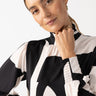 A person with long hair is wearing the On The Spot Blouse Formation by Sanctuary Clothing, featuring a black and white pattern with ruched details on the sleeves. They are looking at the camera with a slight smile, resting their chin on one hand, while gold hoop earrings and rings are visible.