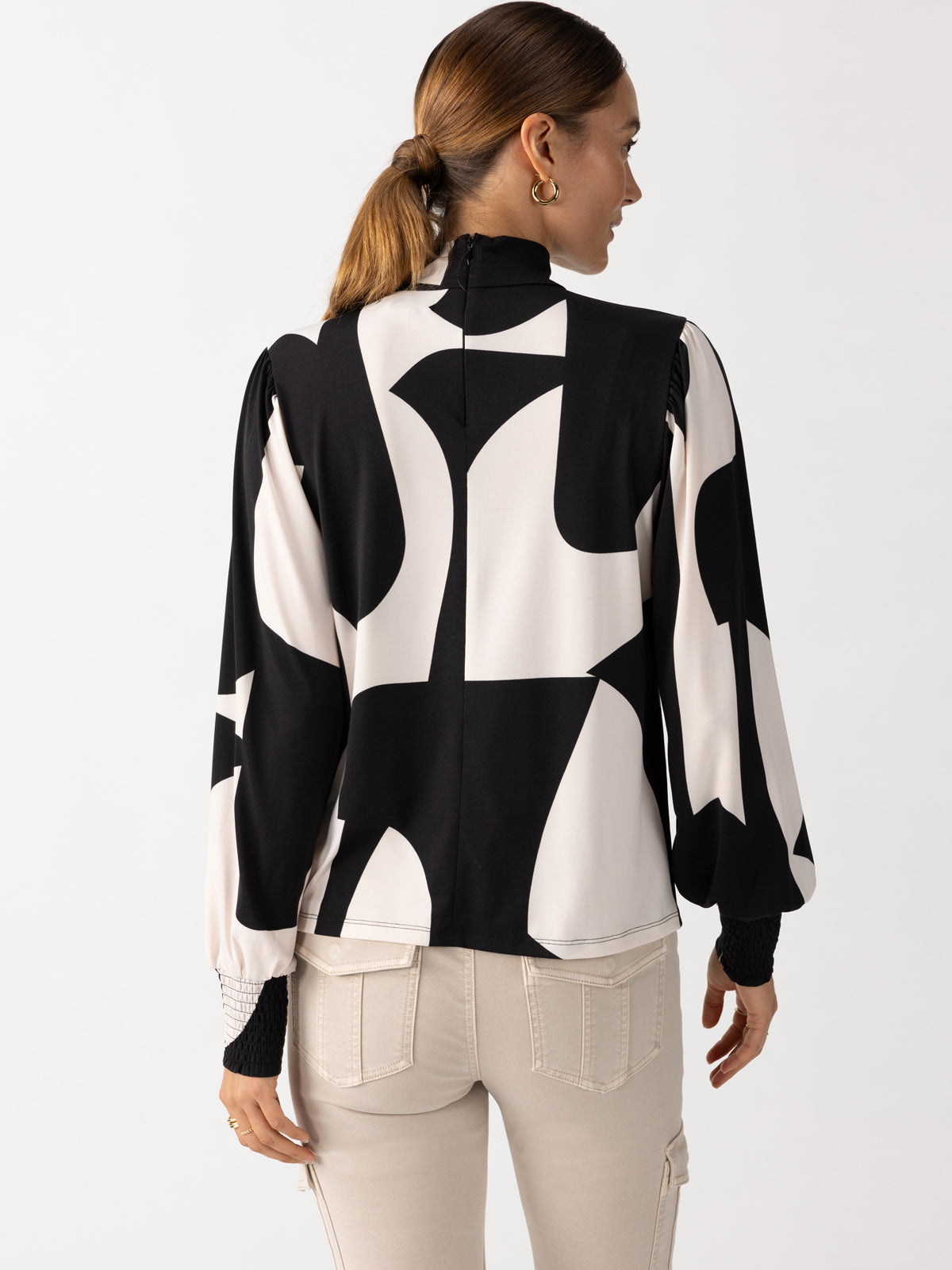 A woman with brown hair in a ponytail is wearing Sanctuary Clothing's On The Spot Blouse Formation, which features a bold black and white geometric pattern and puffed sleeves. She is also dressed in light-colored pants and is facing away, looking slightly to the left.