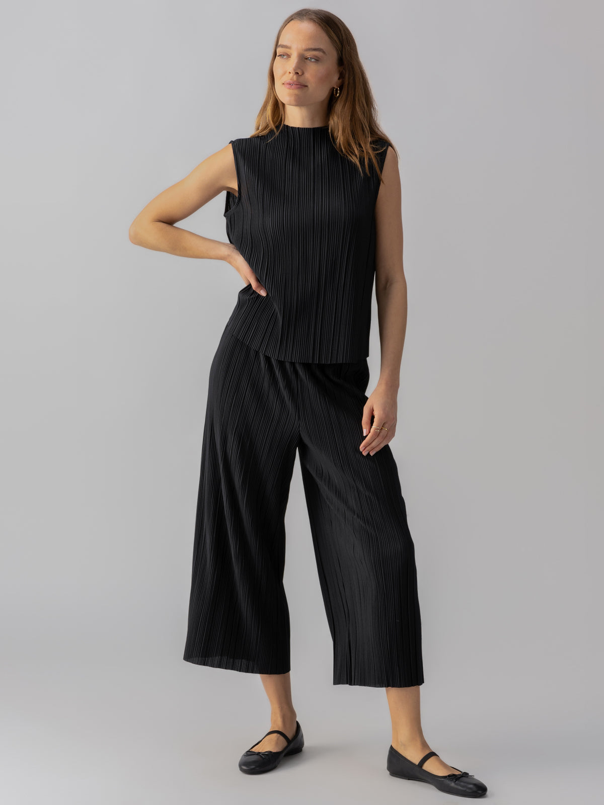 A woman stands against a neutral background, wearing a sleeveless, pleated top paired with the Plisse Culotte Black from Sanctuary Clothing. She has long hair, is wearing minimal makeup, and has a relaxed and confident expression. She completes the outfit with black ballet flats.