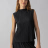 A person with long, light brown hair is wearing the sleeveless Plisse Mock Neck Top Black from Sanctuary Clothing along with matching pleated pants. They are standing against a plain gray background, looking at the camera with a neutral expression.