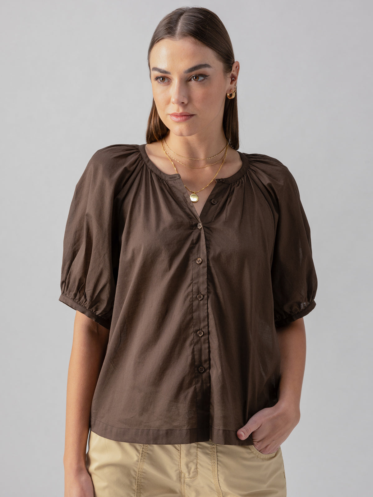 A person with long brown hair and subtle makeup is wearing the Sanctuary Clothing Bubble Sleeve Boho Blouse in Mud Bath, paired with beige pants. They are standing against a plain light gray background, gazing slightly to the side with a calm expression.