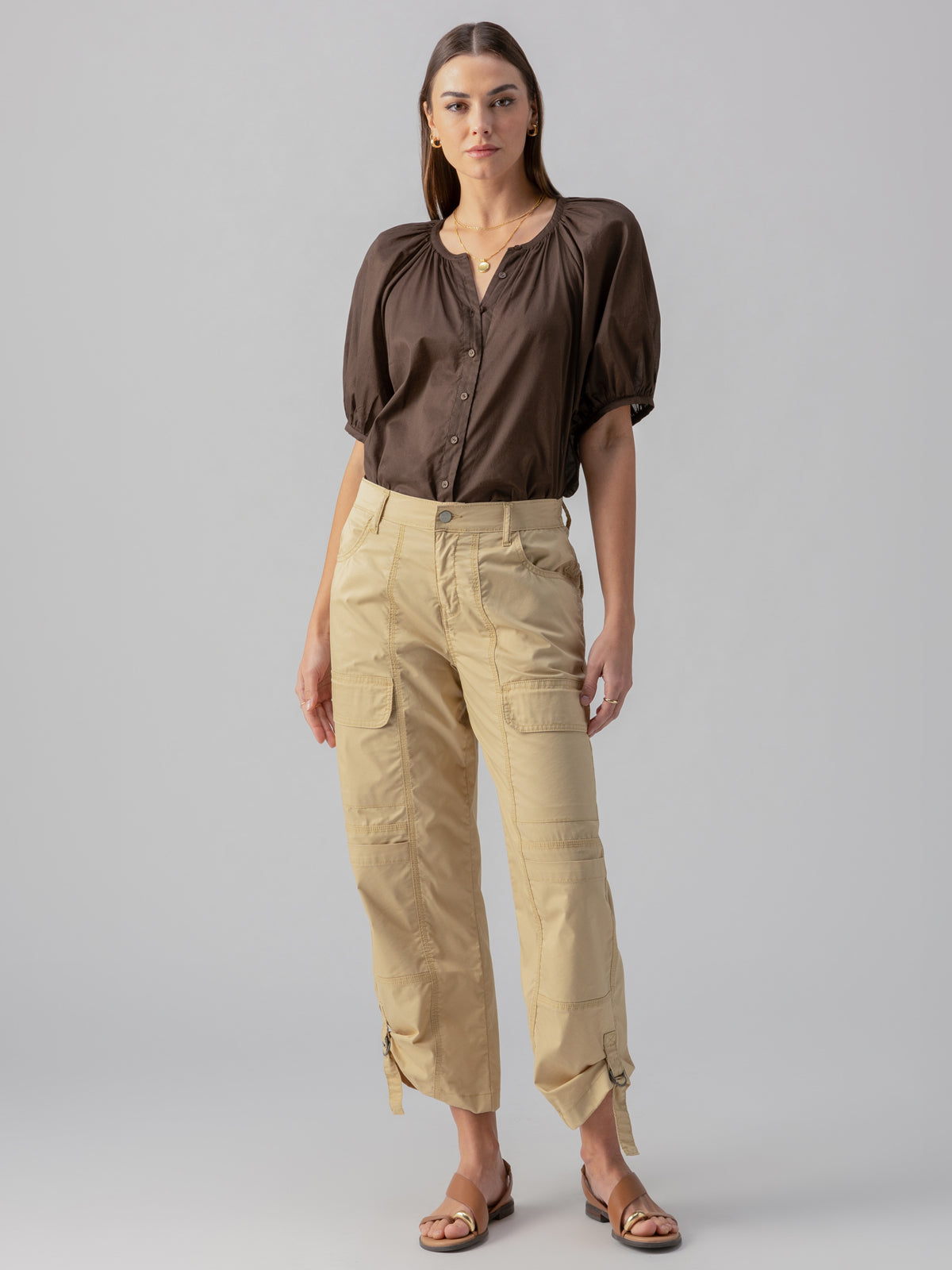 A woman stands against a plain background, wearing the Bubble Sleeve Boho Blouse in Mud Bath by Sanctuary Clothing, light beige cargo pants with multiple pockets and buckles, and brown sandals. She has long, straight hair and minimal jewelry.