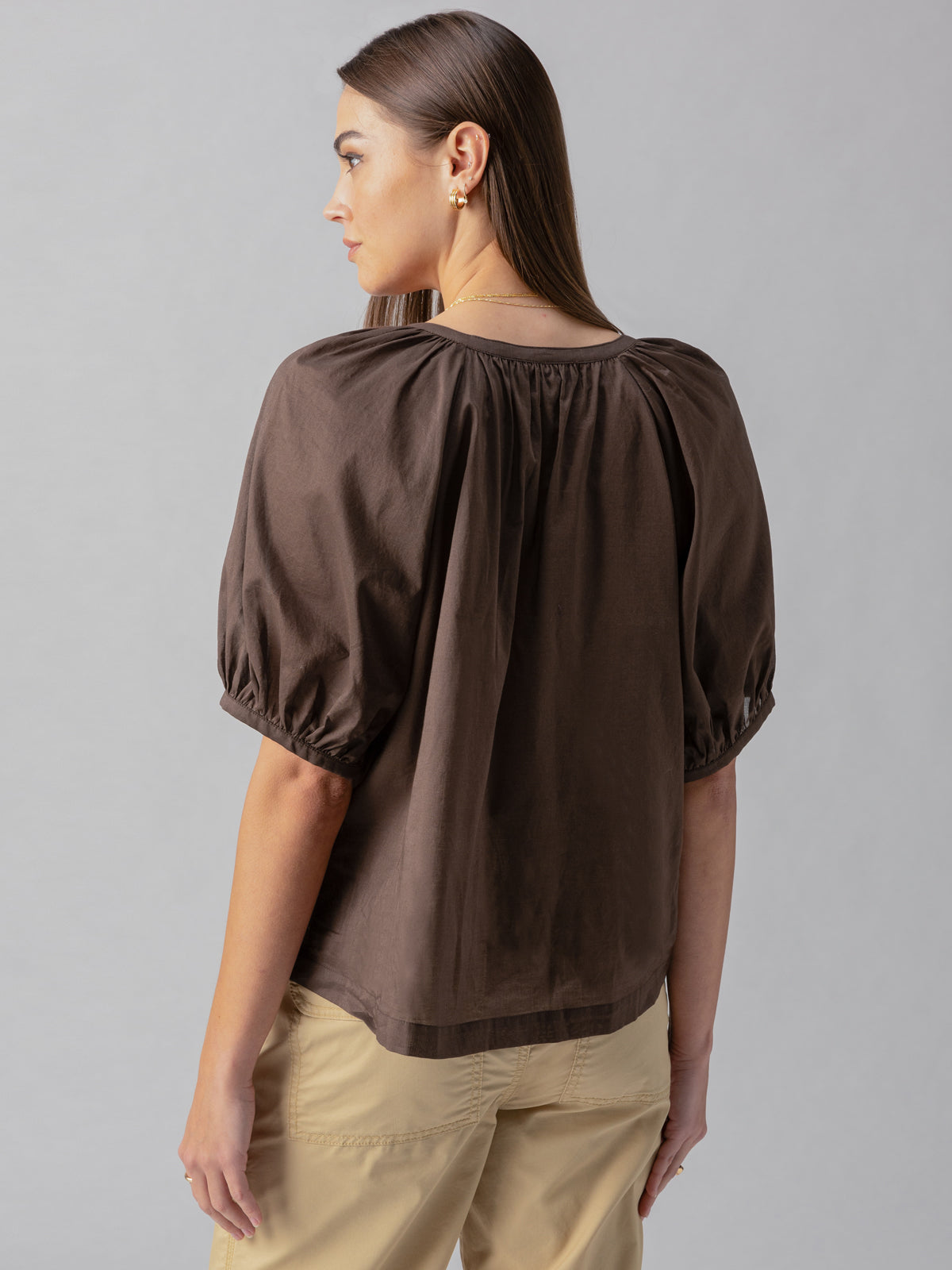 A woman with long brown hair stands facing away, wearing the Sanctuary Clothing Bubble Sleeve Boho Blouse in Mud Bath color with gathered detailing at the shoulders and loose beige pants. The background is neutral.