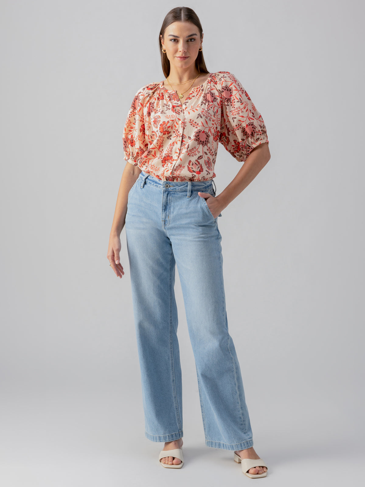 A woman stands confidently with hands in pockets, wearing the Bubble Sleeve Boho Blouse Pacific Light by Sanctuary Clothing and light blue high-waisted jeans. She has brown hair and wears strappy white sandals. The background is plain gray.