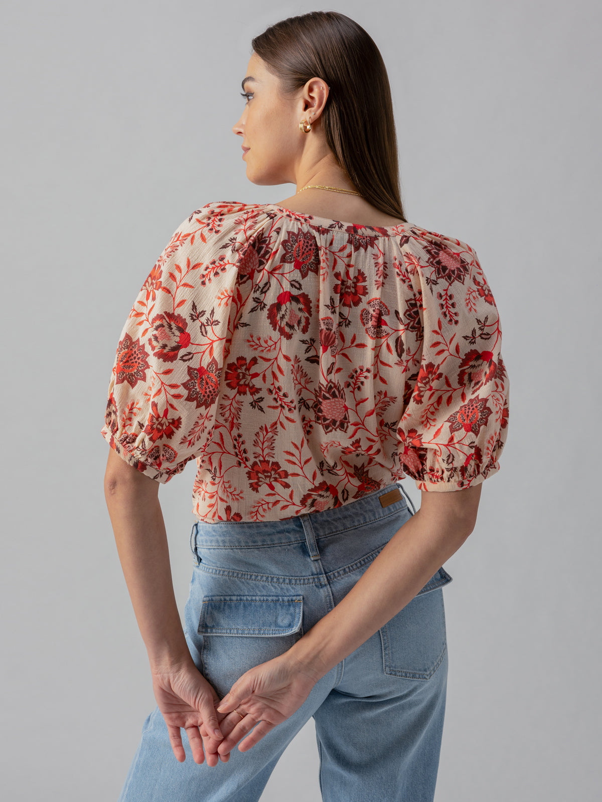 A woman with long brown hair is posing with her back to the camera. She is wearing Sanctuary Clothing’s Bubble Sleeve Boho Blouse Pacific Light, a cream-colored blouse adorned with a red floral pattern, paired with light blue jeans. Her hands are casually clasped behind her back against a plain, light gray background.