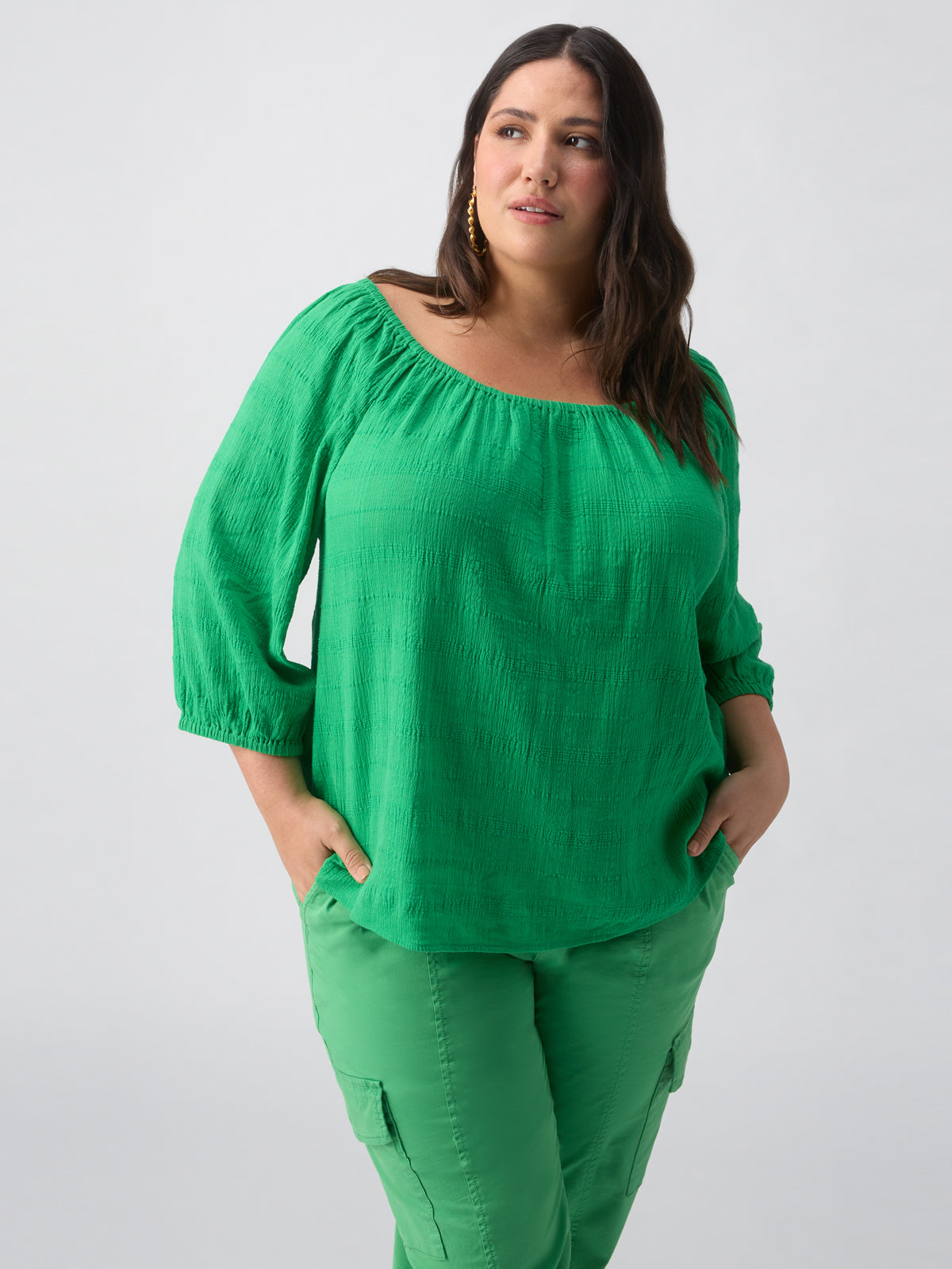 A woman with long dark hair wearing the Beach To Bar Blouse Green Goddess Inclusive Collection by Sanctuary Clothing, tucked into green cargo pants, stands against a plain white background. She is looking slightly to the side with her hands in her pockets.