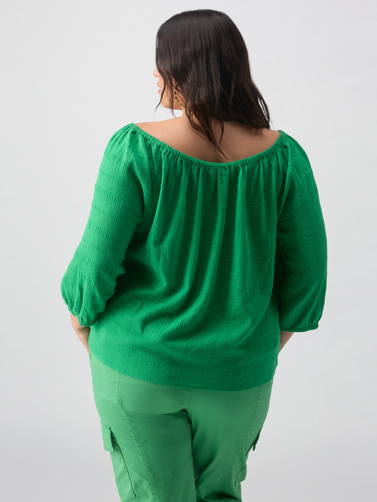 A person with long dark hair is facing away from the camera, wearing a Sanctuary Clothing Beach To Bar Blouse Green Goddess Inclusive Collection, along with green cargo pants. The background is plain and light-colored.