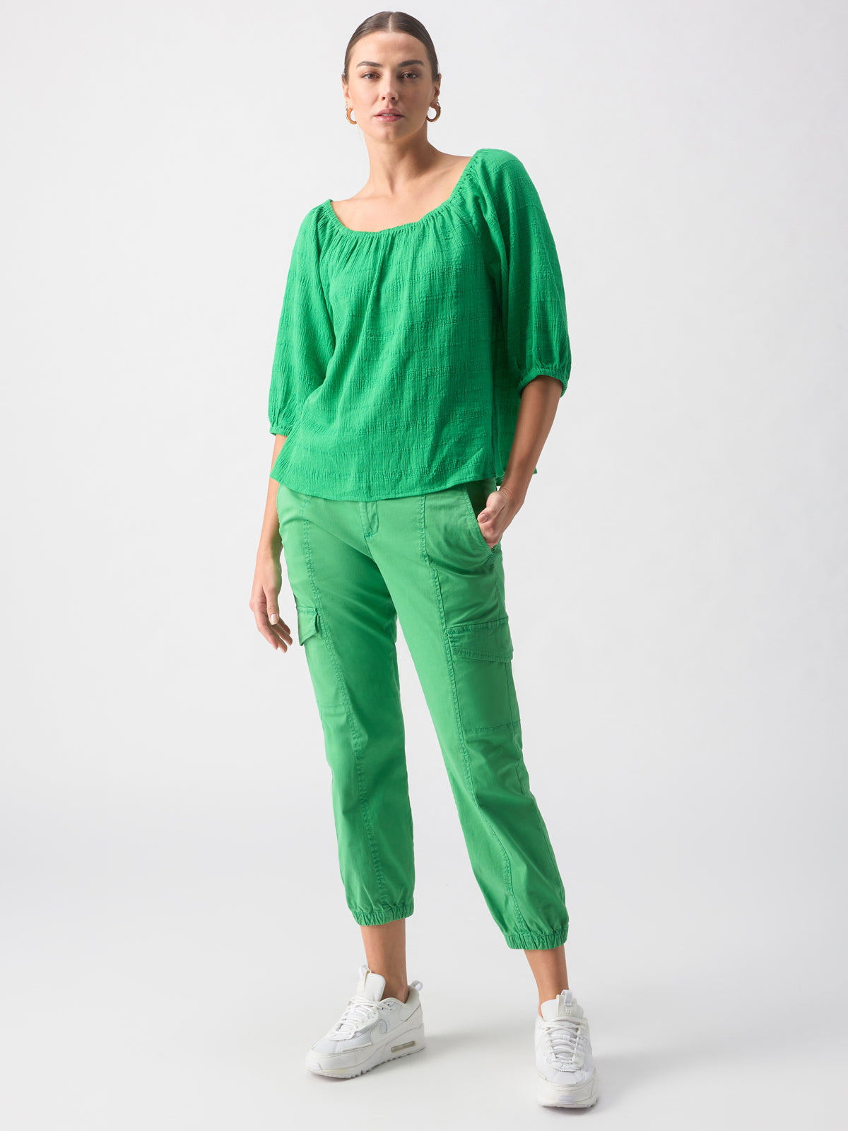 A person stands confidently against a plain background. They are dressed in a loose, green three-quarter sleeve top called the "Beach To Bar Blouse Green Goddess" by Sanctuary Clothing, paired with green cargo pants that gather at the ankles. They are also wearing white sneakers and large hoop earrings.