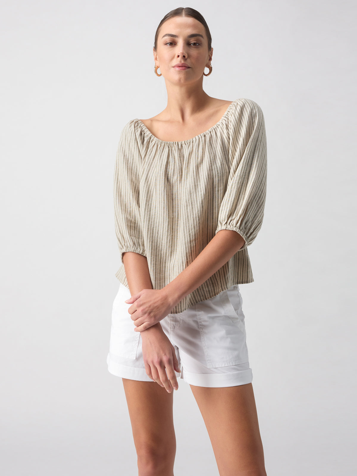 A person stands against a plain background wearing the Beach To Bar Blouse Eco Olive Stripe by Sanctuary Clothing, paired with white shorts. The blouse is beige with stripes and features an off-shoulder design and three-quarter sleeves. They have their hands crossed casually in front of them and are looking directly at the camera with a neutral expression.