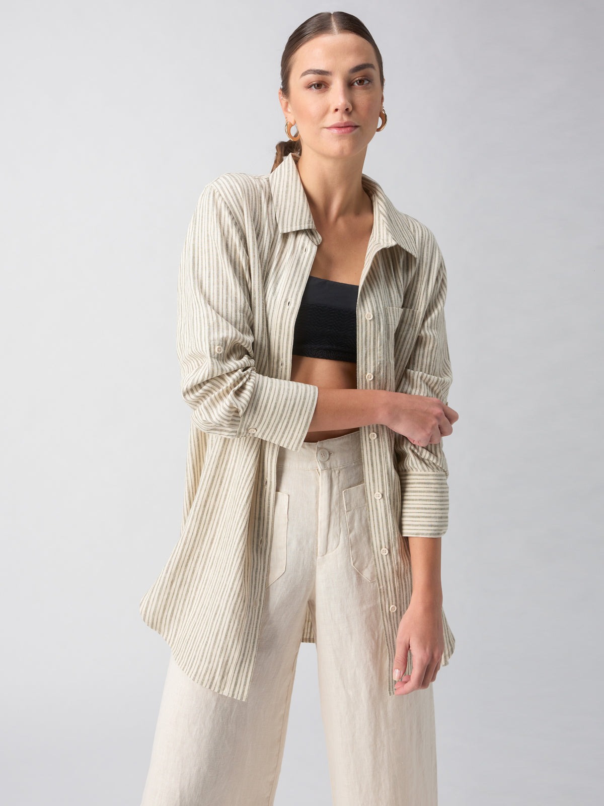 A person poses confidently against a plain gray background. They are wearing Sanctuary Clothing's Long Line Pocket Shirt in Eco Olive Stripe over a black crop top, paired with light beige pants. The person's hair is pulled back, and they are accessorized with hoop earrings.