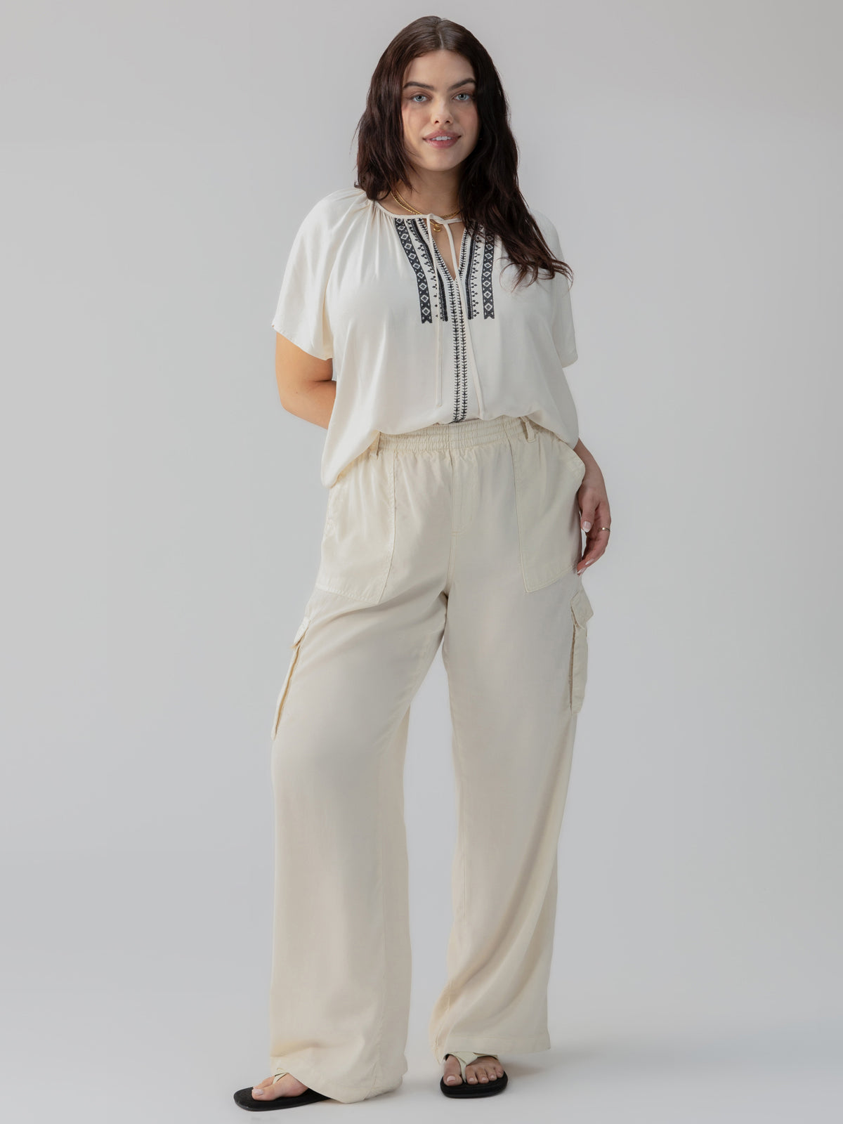 A woman stands confidently wearing the Sanctuary Clothing's Golden Dream Top Birch from the Inclusive Collection, a loose-fitting white blouse with black embroidery on the front. She pairs it with flowy beige pants featuring cargo pockets. Her long brown hair cascades down as she steps into black sandals against a plain light gray background.