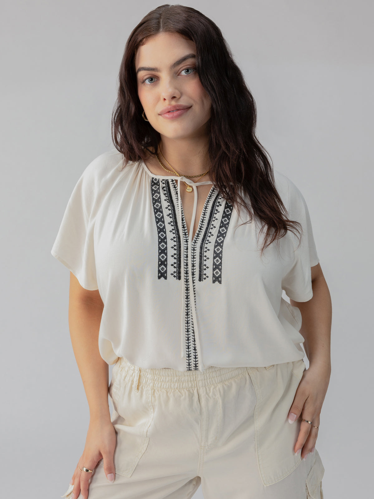 A person wearing the Golden Dream Top Birch from Sanctuary Clothing's Inclusive Collection, featuring black embroidered patterns along the neckline and front, paired with light-colored shorts. They are standing in front of a neutral background, with their hair down and a gentle expression on their face.