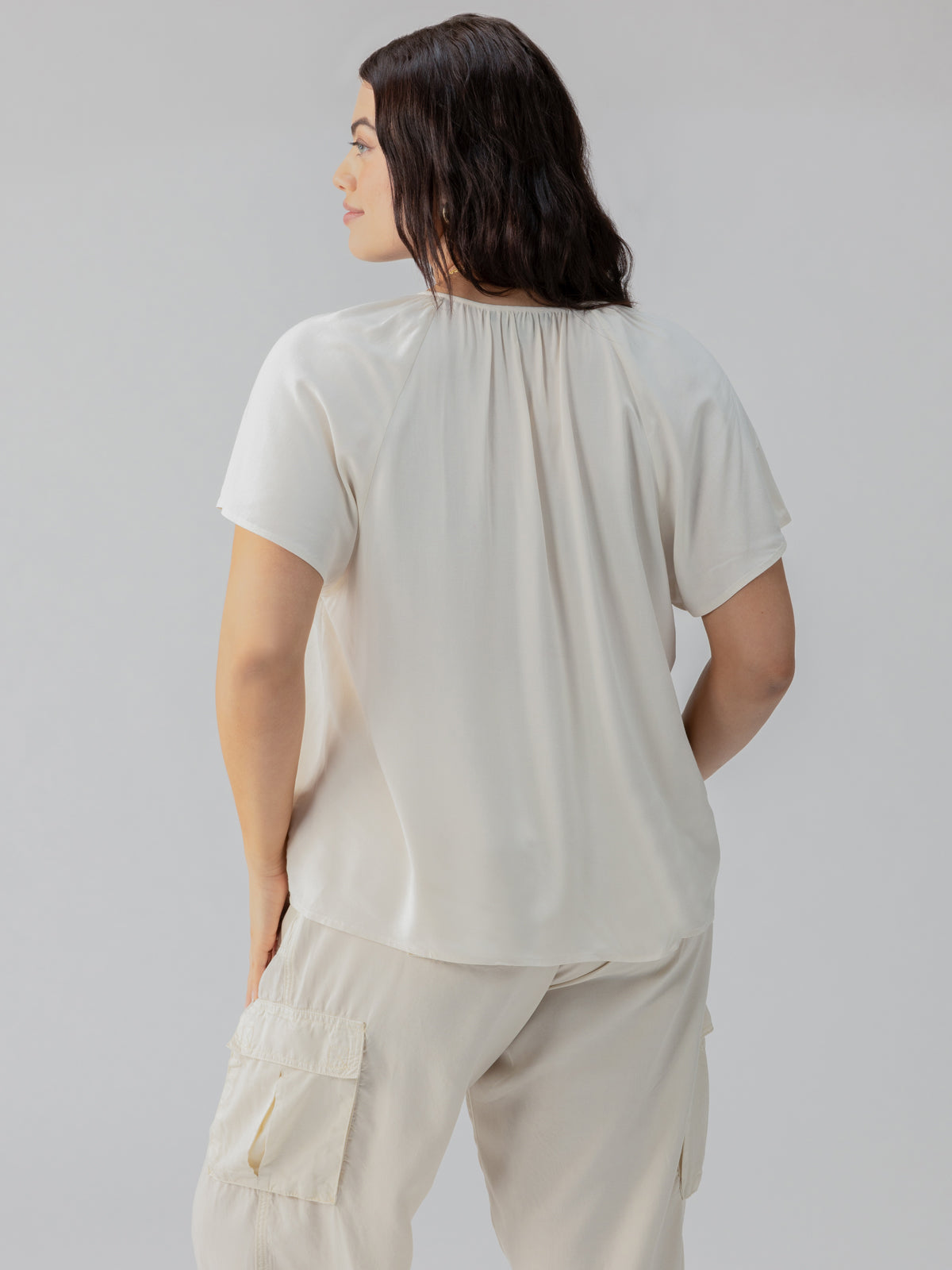 A person with shoulder-length dark hair faces sideways, wearing the Golden Dream Top Birch Inclusive Collection from Sanctuary Clothing. They are also donning matching light-colored pants with cargo pockets, set against a simple light gray background.