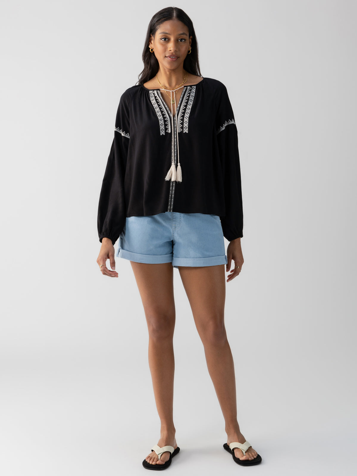 A person with long hair stands against a plain background, wearing a Sanctuary Clothing Embroidered Blouse Black with white embroidery, light blue shorts, and white sandals. The blouse features dangling tassels, and the person has a neutral expression and relaxed posture.