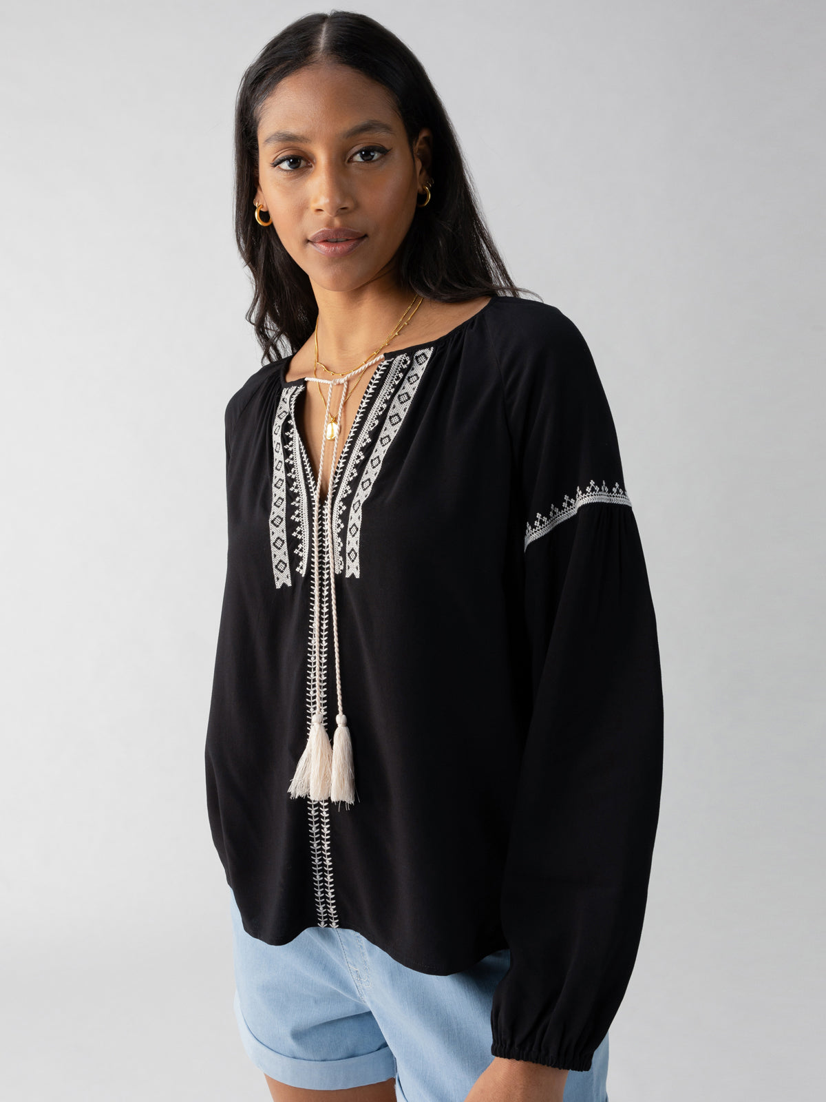 A woman with long dark hair is wearing the Embroidered Blouse Black by Sanctuary Clothing, which features white embroidered patterns along the neckline and sleeves, as well as tassels at the neck. She is also wearing light blue shorts and stands with a neutral expression against a plain, light gray background.