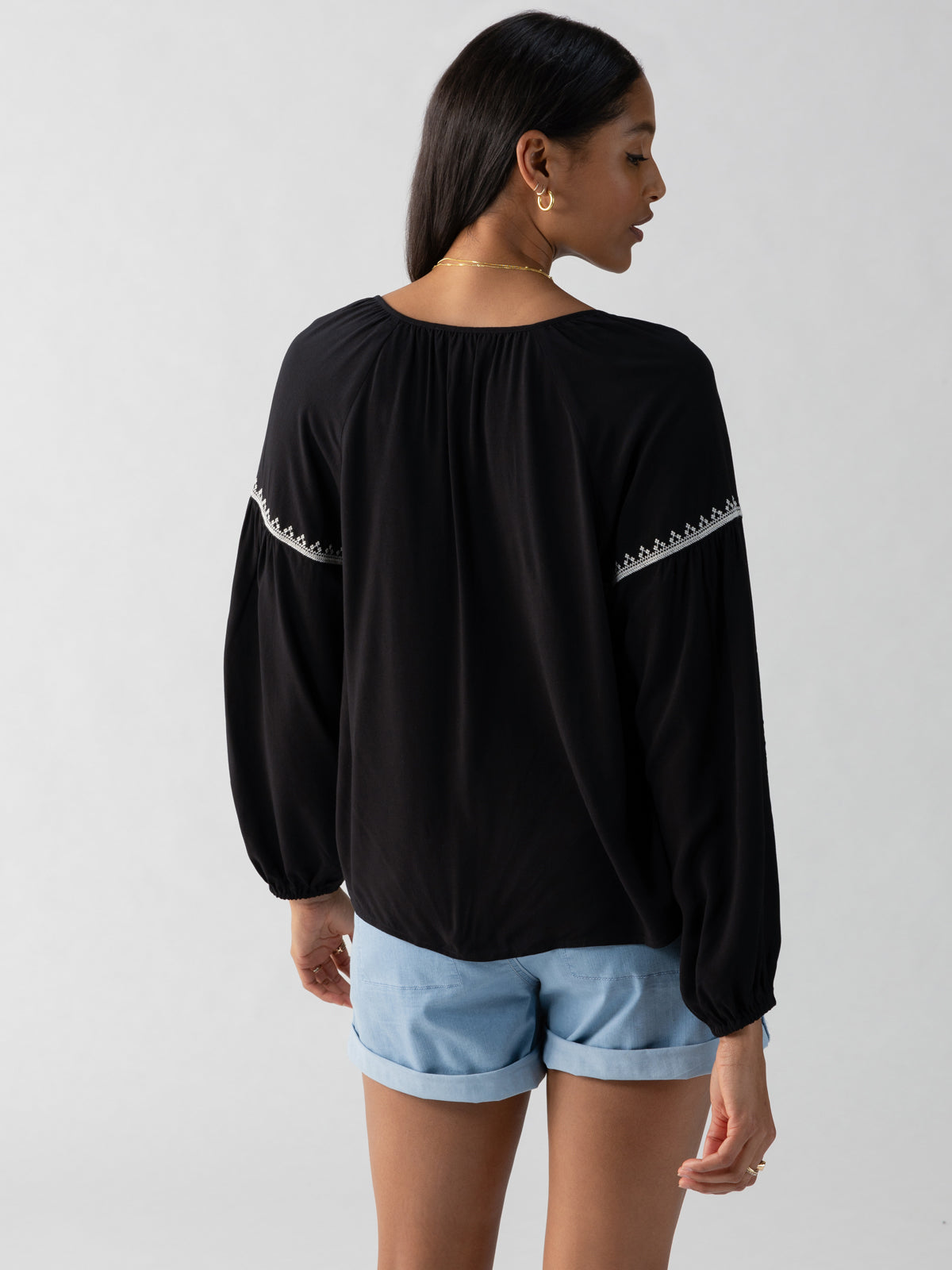 A woman stands facing away, looking to the side. She has long dark hair and is wearing the Embroidered Blouse Black by Sanctuary Clothing, featuring white embroidery along the shoulder line. She pairs it with light blue shorts against a plain, light-colored background.