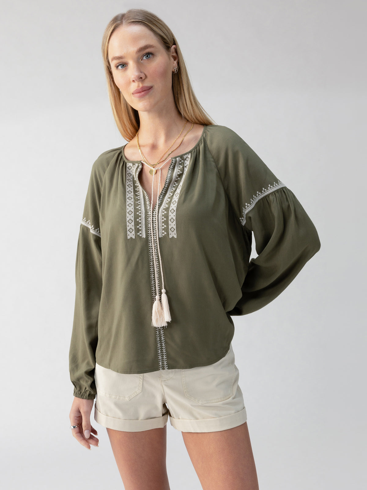 A woman with long blonde hair is wearing the Embroidered Blouse in Burnt Olive by Sanctuary Clothing, featuring white embroidered details and cream-colored tassels. She pairs it with light beige shorts and stands against a plain light gray background.