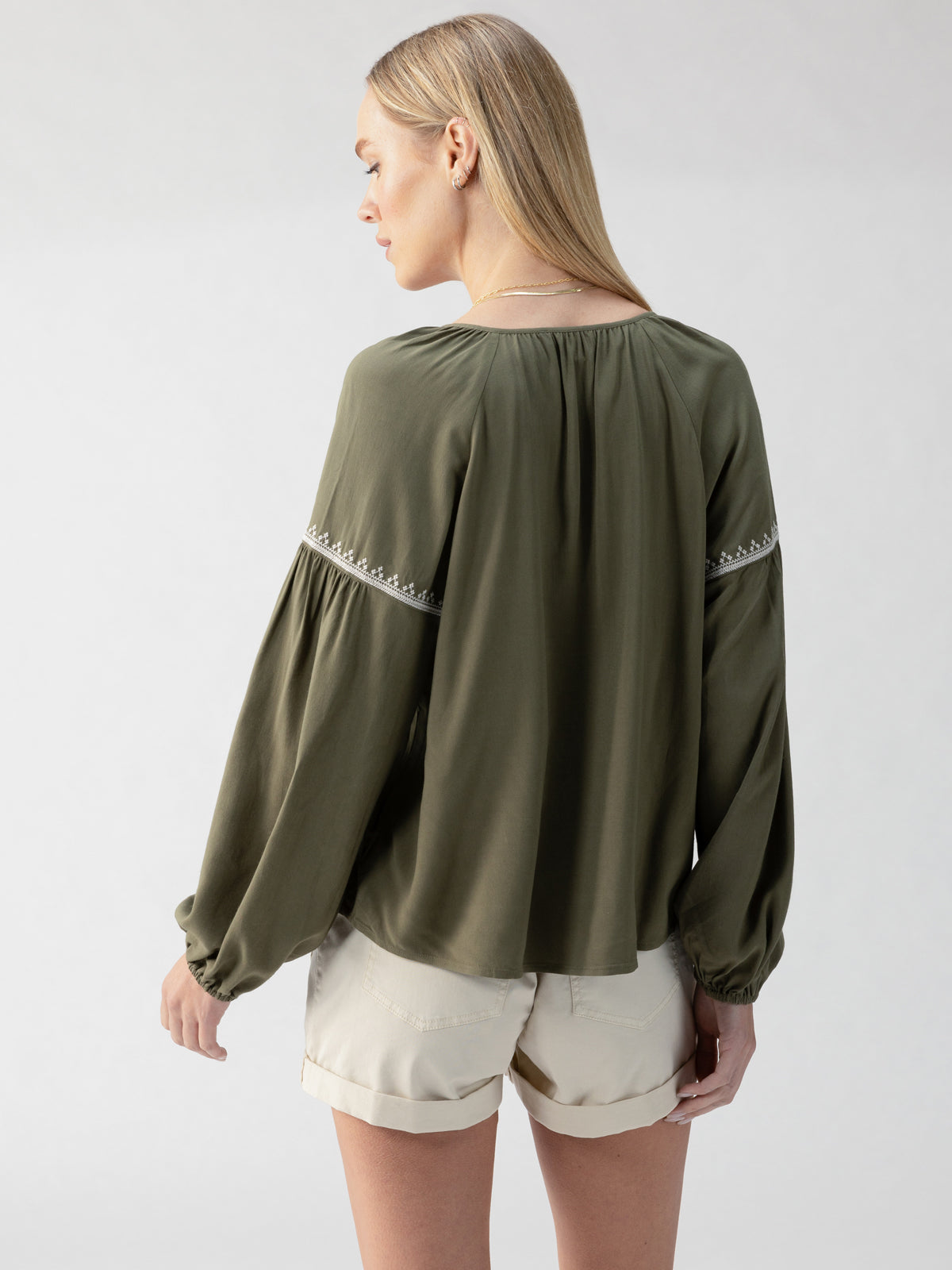 A woman with long blonde hair stands with her back turned, wearing the "Embroidered Blouse Burnt Olive" by Sanctuary Clothing, paired with beige shorts. The blouse features flowy sleeves adorned with intricate embroidery, and she is in a neutral, well-lit indoor setting.