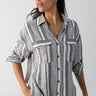 A person wearing the Sanctuary Clothing Pocket Shirt Varigated Stripe, a long-sleeved, button-down shirt with vertical gray and white stripes. They have their hands in their pockets and are looking to the side. They have long, dark hair and are accessorized with gold necklaces and earrings.