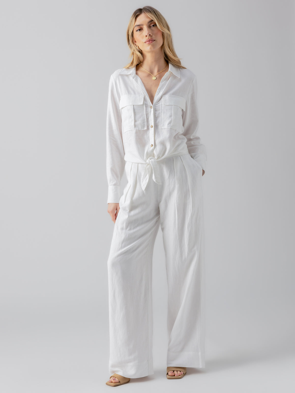 A person with shoulder-length hair stands against a neutral background, wearing a Utility Pocket Shirt White by Sanctuary Clothing. They pair it with matching high-waisted, wide-leg pants and have one hand in their pocket while wearing sandals.