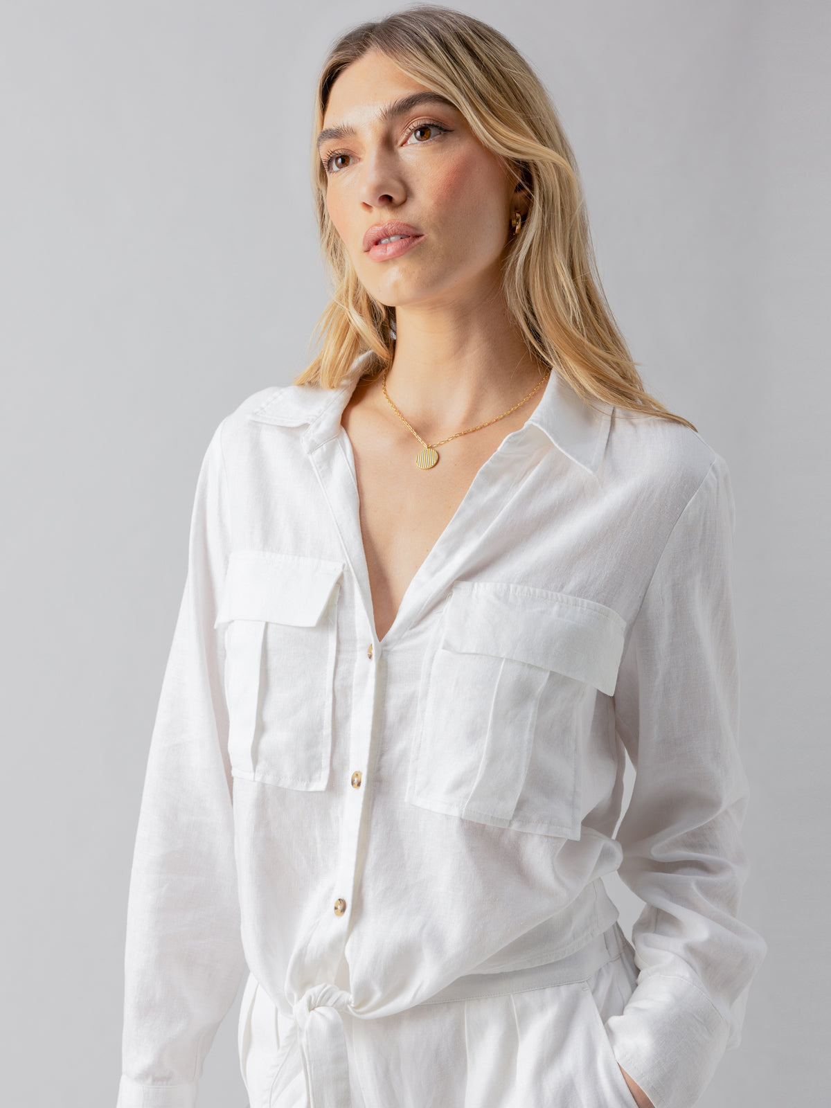 A woman with long blonde hair, adorned in the Utility Pocket Shirt White by Sanctuary Clothing, accessorized with a gold necklace, stands against a plain light gray background. Her calm expression and slight sideways glance complete the serene imagery.