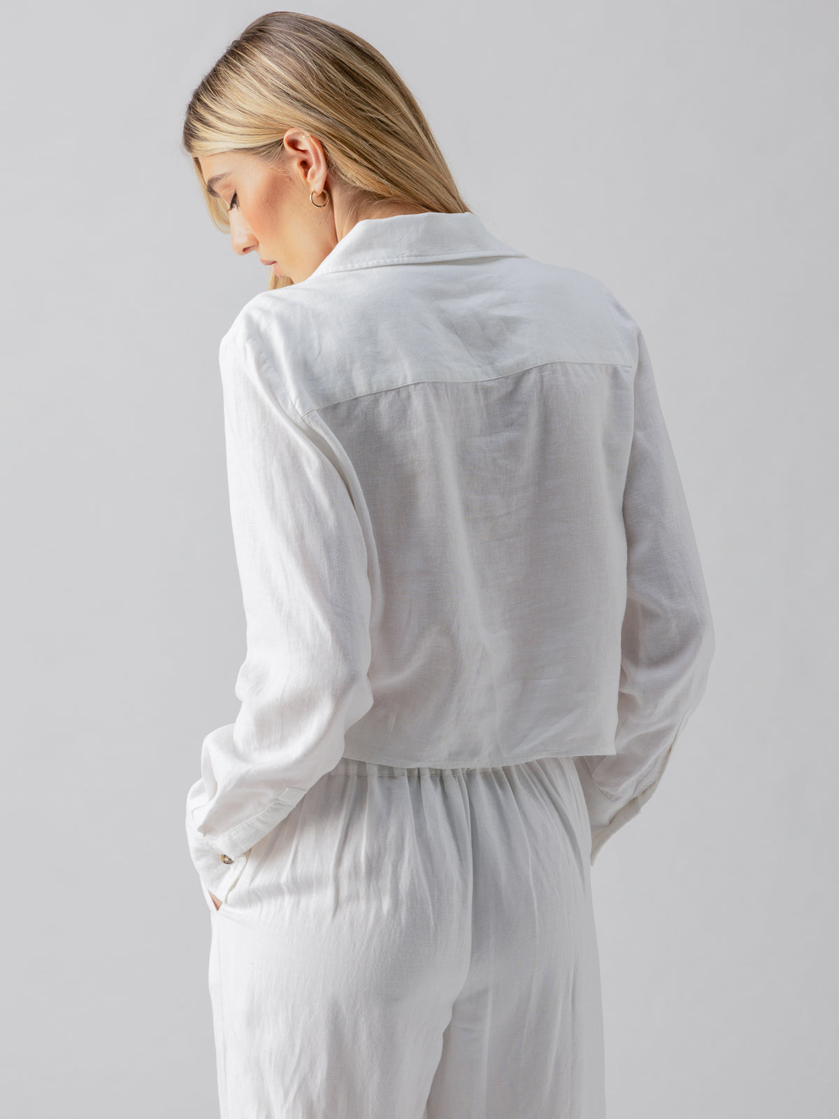 A person with blonde hair is seen from behind, wearing the Sanctuary Clothing Utility Pocket Shirt in white and matching pants. Their gaze is directed downward, with one hand tucked into a pocket, creating a casual pose against a plain background.